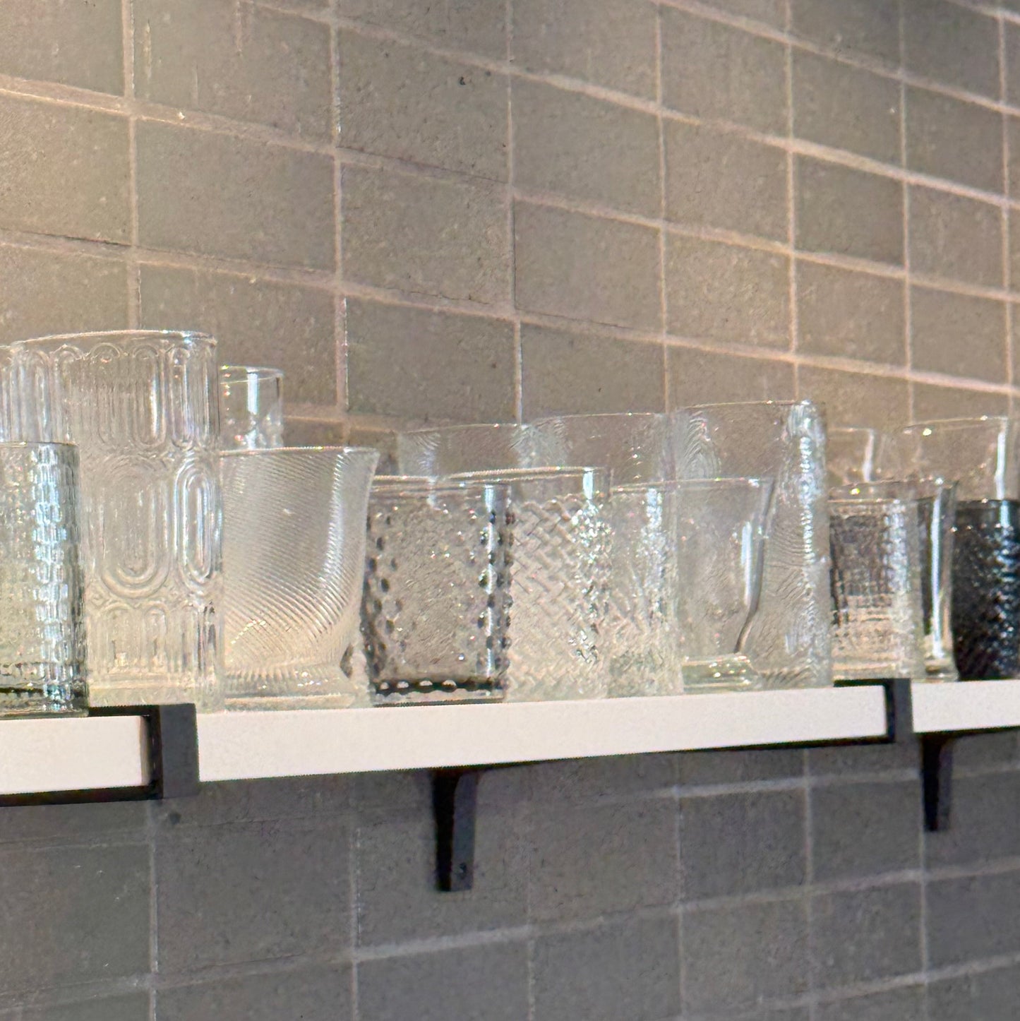 Set of clear glass tumblers on a shelf against a tiled wall.
