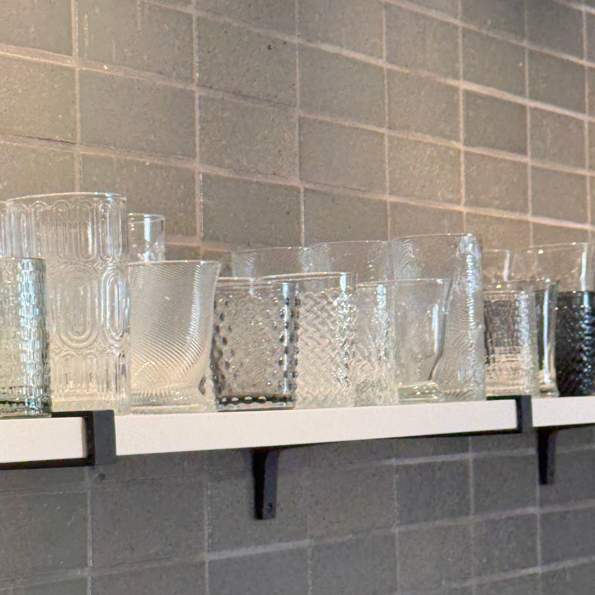 Set of clear glass tumblers on a shelf against a tiled wall.