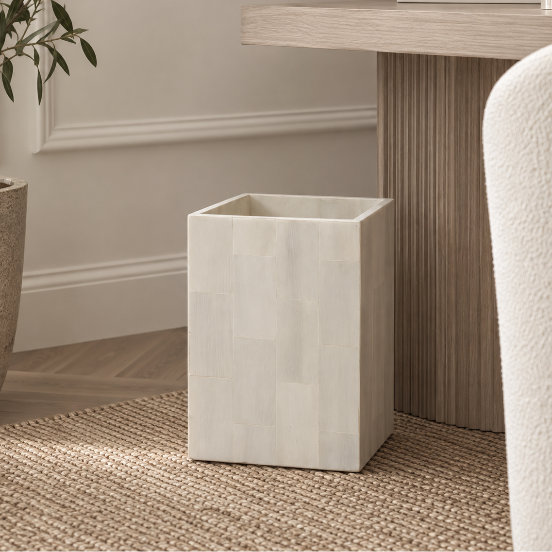 A rectangular off-white bone/shell-look wastebasket with a patchwork texture, styled in a modern living room setting next to a wooden console table and textured rug.