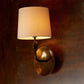Wall-mounted lamp with a gold fixture and beige shade on a dark brown wall.