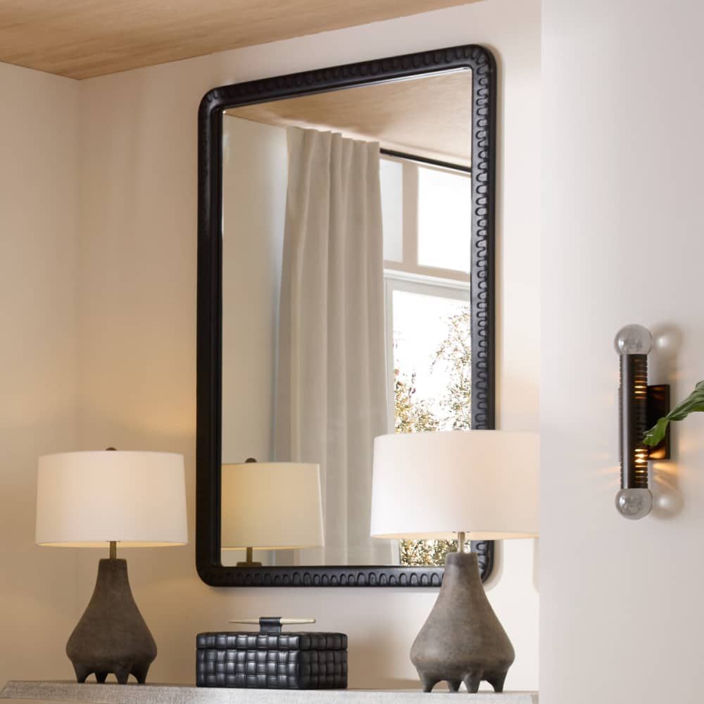 Decorative mirror with black frame on a wall above a table with lamps.