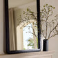Black-framed mirror on a wall with decorative branches and a vase.