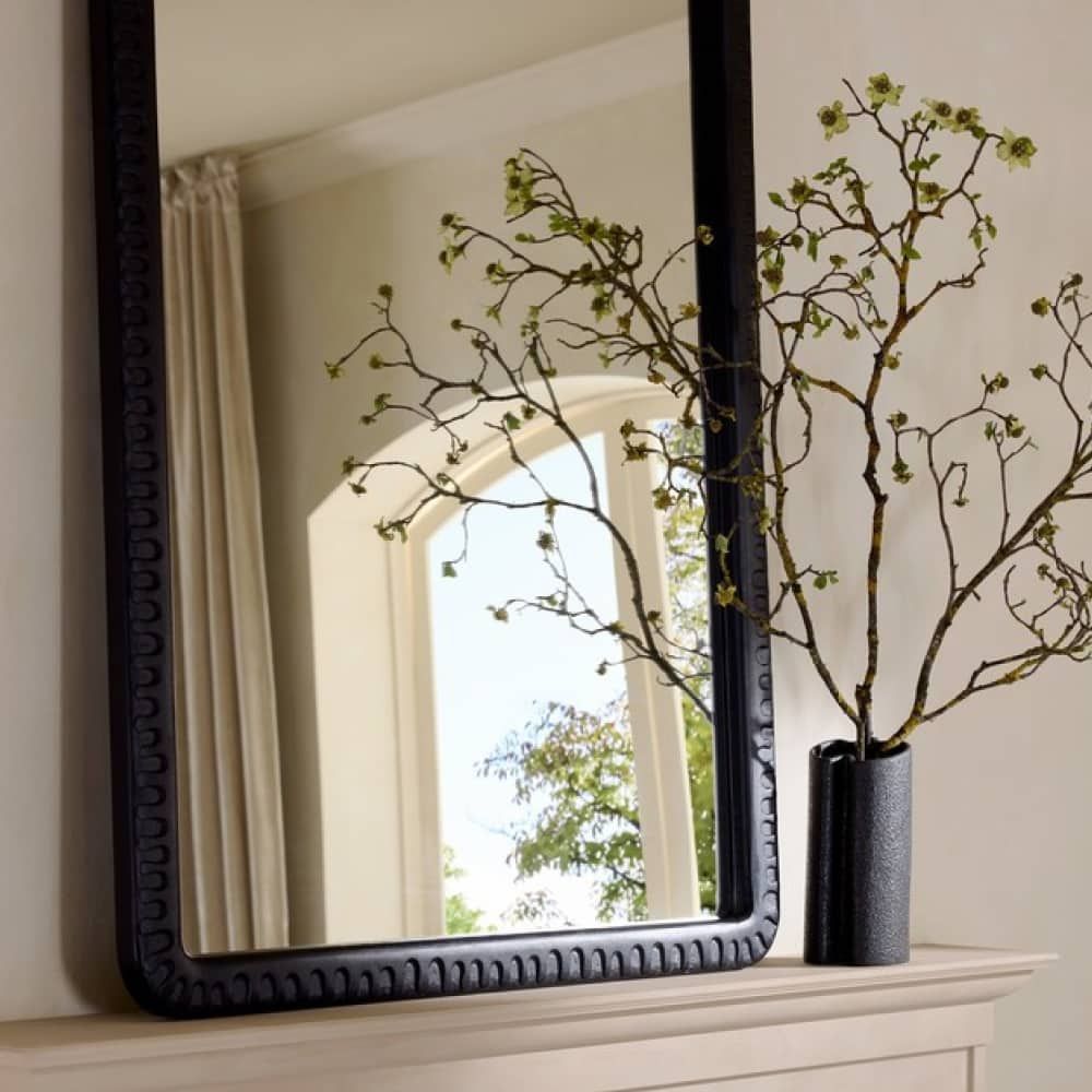 Black-framed mirror on a wall with decorative branches and a vase.