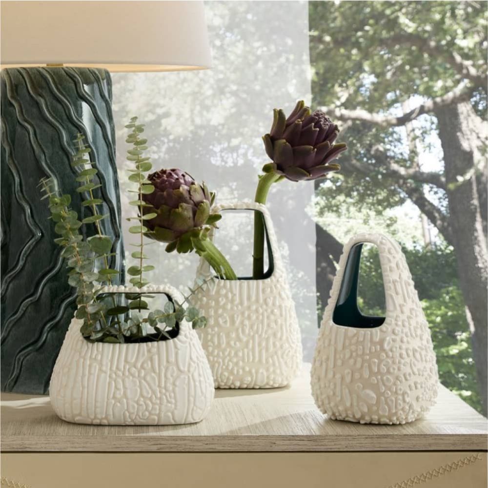Three textured white vases with plants on a surface near a window with a view of trees.