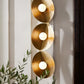 Gold circular wall light fixture with three illuminated bulbs on a neutral background.