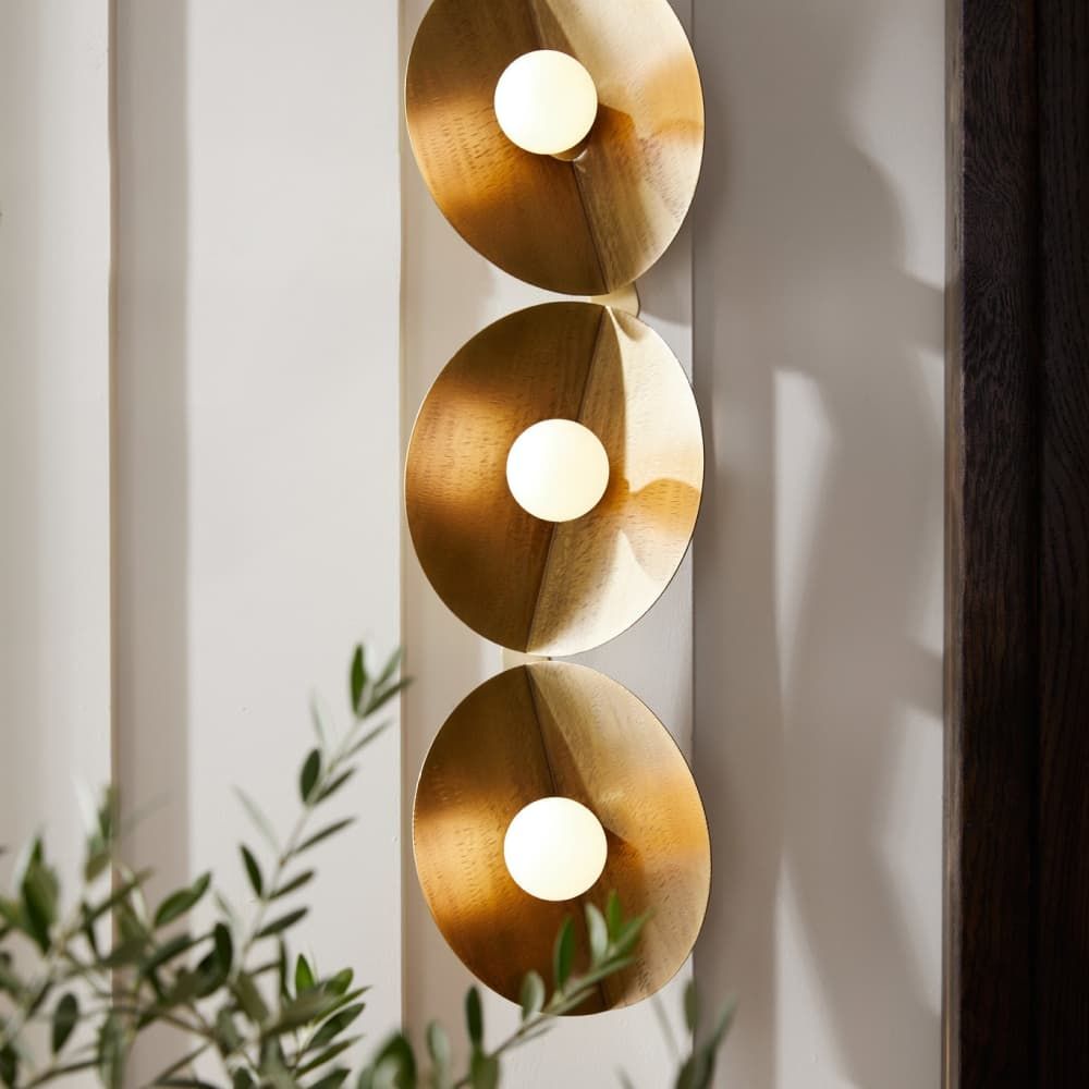 Gold circular wall light fixture with three illuminated bulbs on a neutral background.