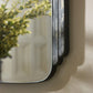 Rectangular mirror with black frame on a textured wall with a plant reflection.
