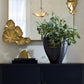 Decorative setup with a black vase, gold leaf sculpture, and green plant on a dark surface.