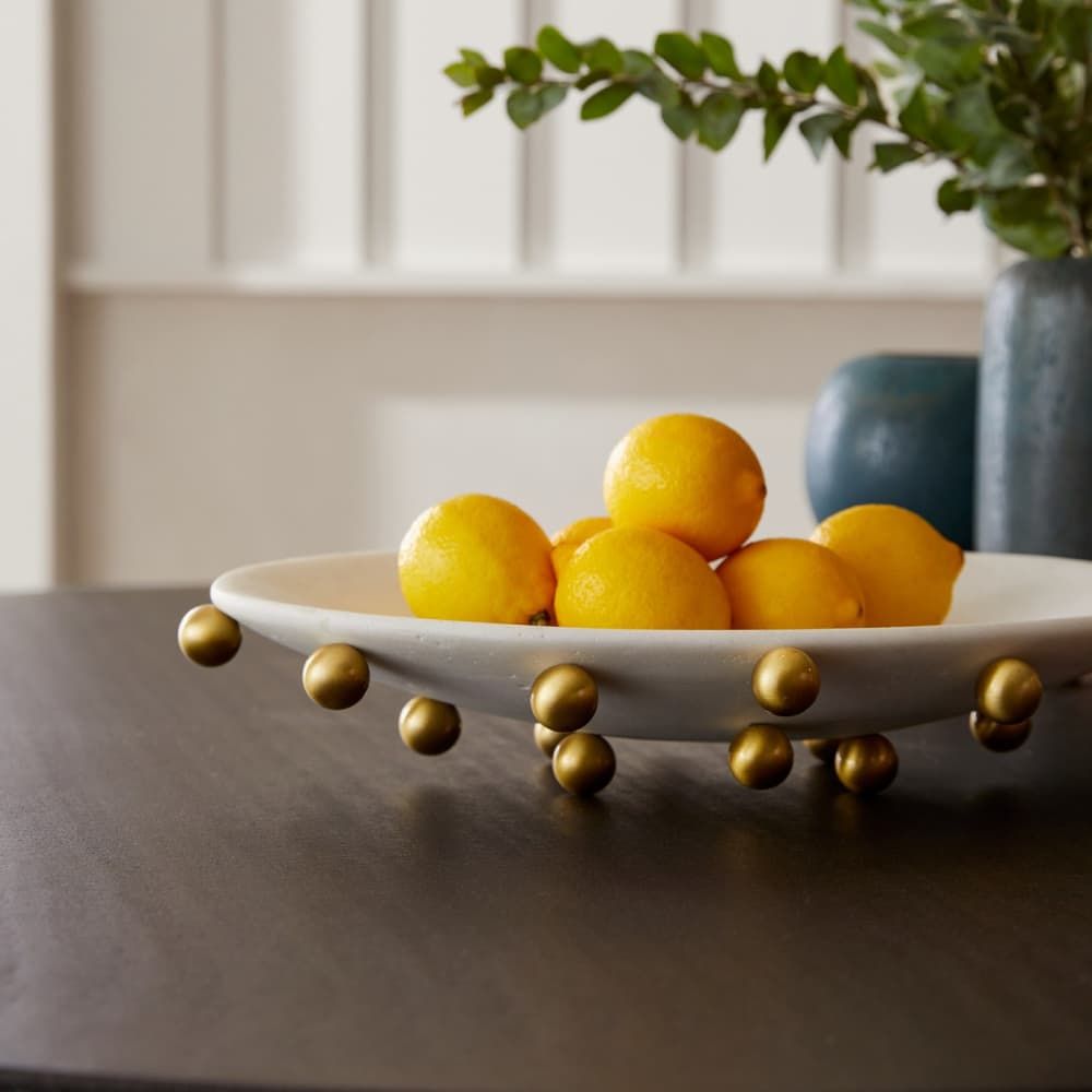 White bowl with gold legs containing lemons on a table with a blurred background