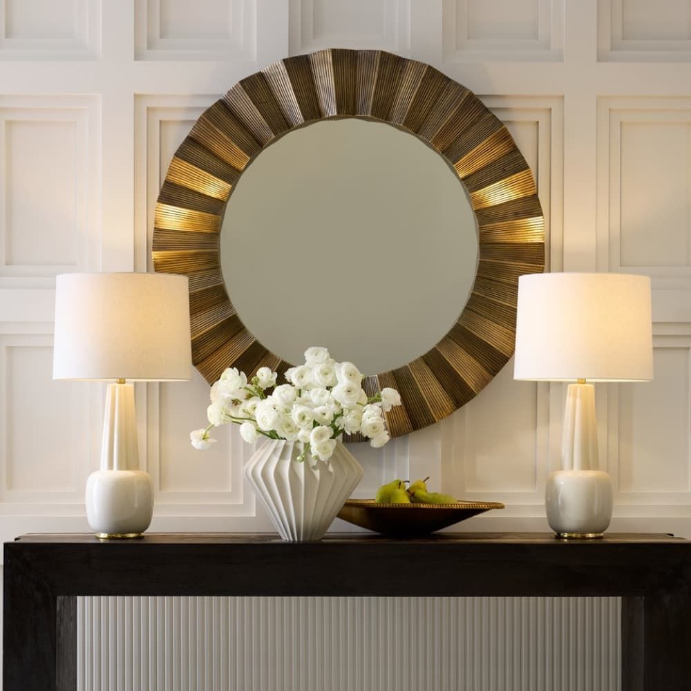 Decorative round mirror with a wooden frame on a wall above a console table with lamps and flowers.