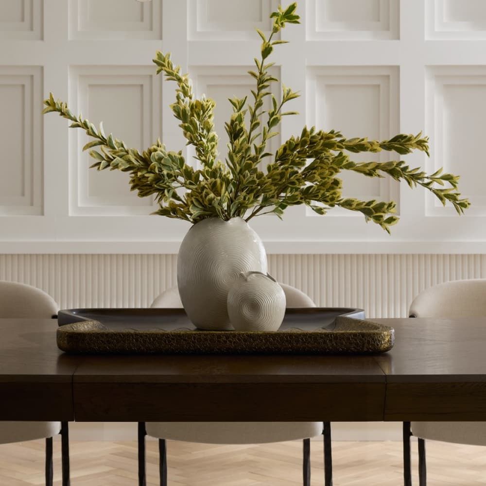 Decorative vase with green branches on a wooden table in a room with white paneled walls.