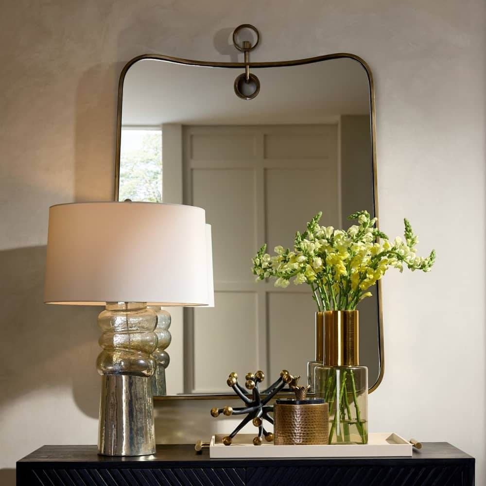 Decorative table with lamp, mirror, and vase of flowers against a neutral wall.