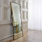 Decorative floor mirror with wavy gold frame in a room with white walls and wooden flooring.