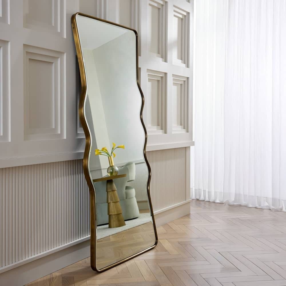 Decorative floor mirror with wavy gold frame in a room with white walls and wooden flooring.