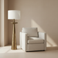 A modern floor lamp with a tapered brushed brass base and a white drum shade stands beside a light beige upholstered armchair in a minimalist room with warm beige walls and soft sunlight shadows.