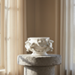 A white ceramic decorative bowl with sculpted floral and scroll motifs, displayed on a round stone pedestal in a softly lit interior with beige walls and sheer curtains.
