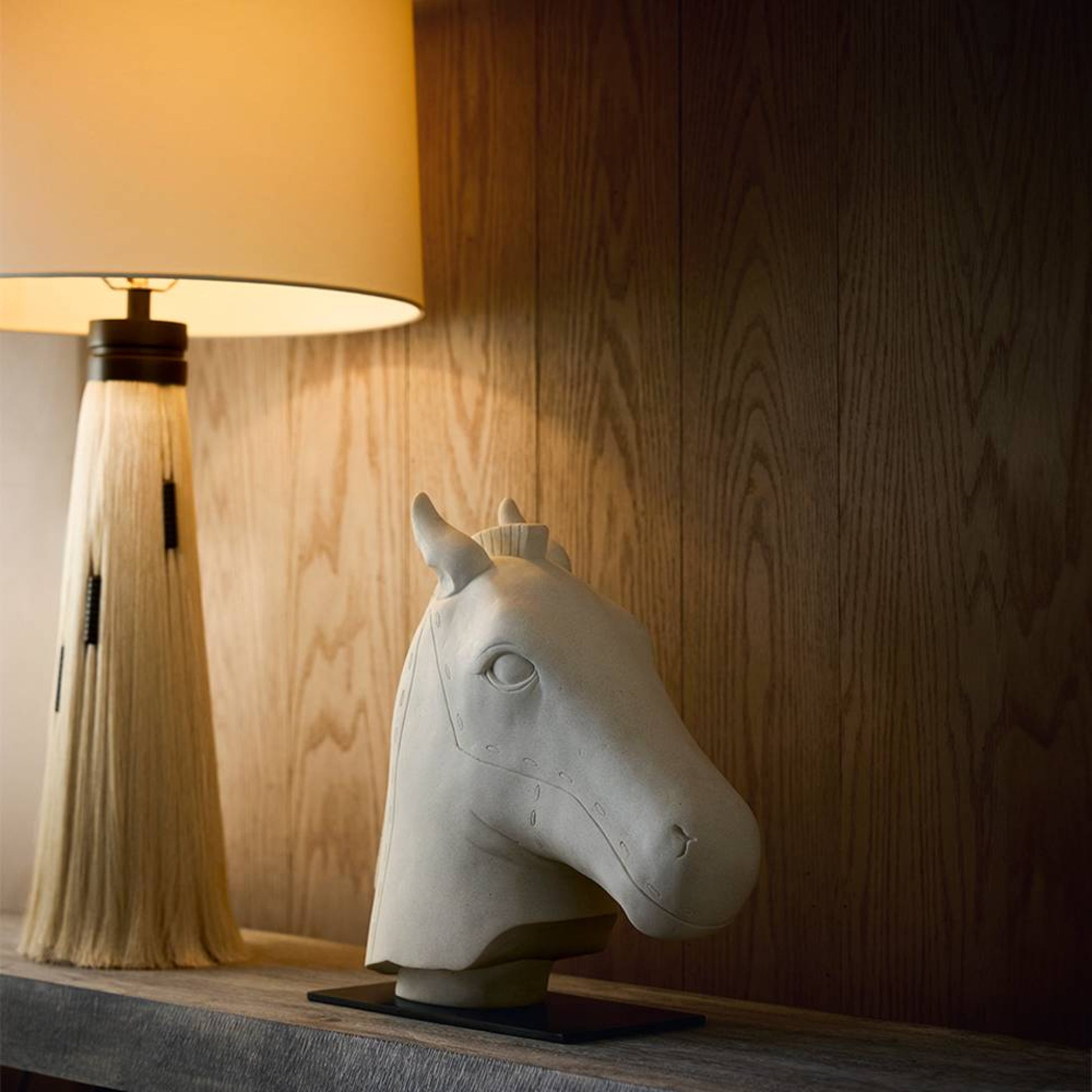 A sculptural horse head table lamp in matte off-white sits on a wooden surface beside a lit table lamp with a beige shade and fringed base against a wood-paneled wall.