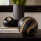 Decorative black and gold sphere on a surface with books and a vase in the background.