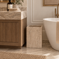A square stone trash can in a warm beige stone finish stands on a tiled bathroom floor between a wooden vanity and a white freestanding bathtub.