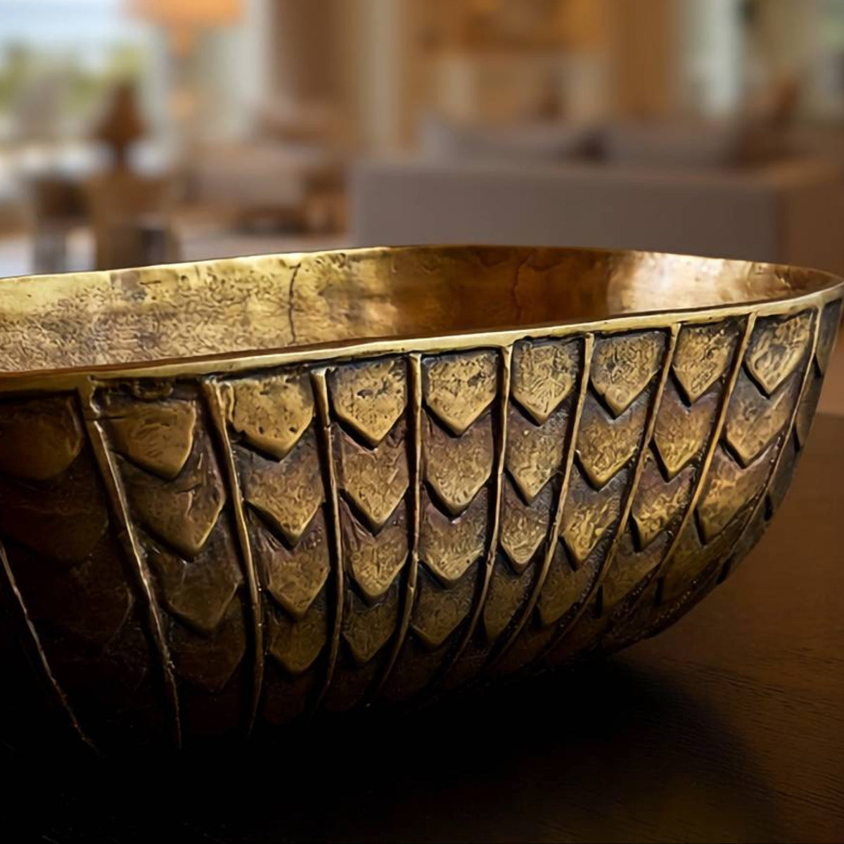 A large decorative metallic bowl with an embossed textured pattern, shown in a warm, blurred interior setting.