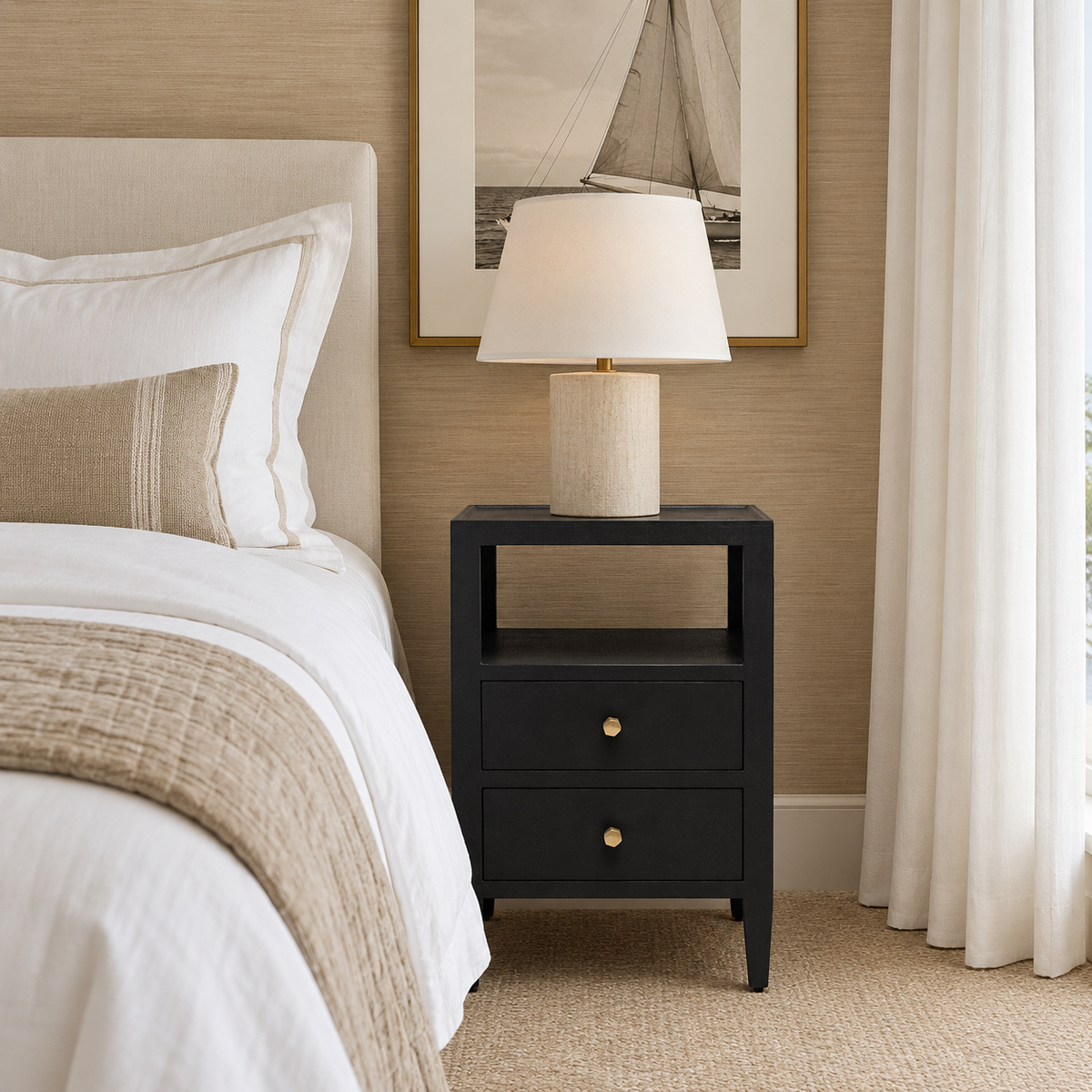 A modern black nightstand with two drawers and brass knobs, styled in a bedroom next to a bed with white linens and a textured beige throw blanket. A table lamp with a beige textured base and white drum shade sits on the nightstand.