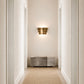 A modern three-tiered bronze wall sconce mounted on a beige wall in a hallway, casting a warm glow above a gray upholstered bench.