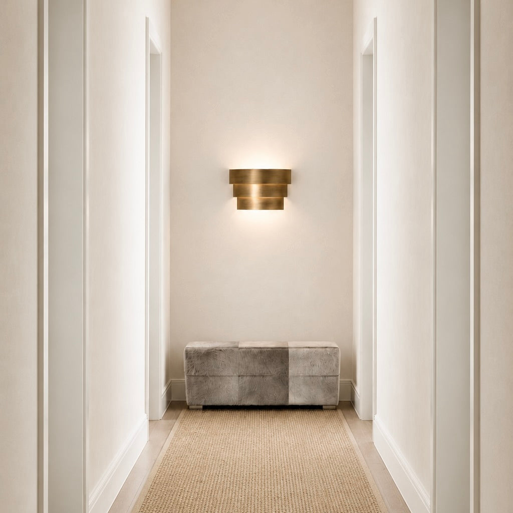 A modern three-tiered bronze wall sconce mounted on a beige wall in a hallway, casting a warm glow above a gray upholstered bench.