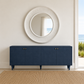 A navy blue sideboard cabinet with four doors and small brass knobs, styled in a coastal interior beneath a large round white mirror. A window on the right shows a view of the ocean, and a woven jute rug covers the floor.