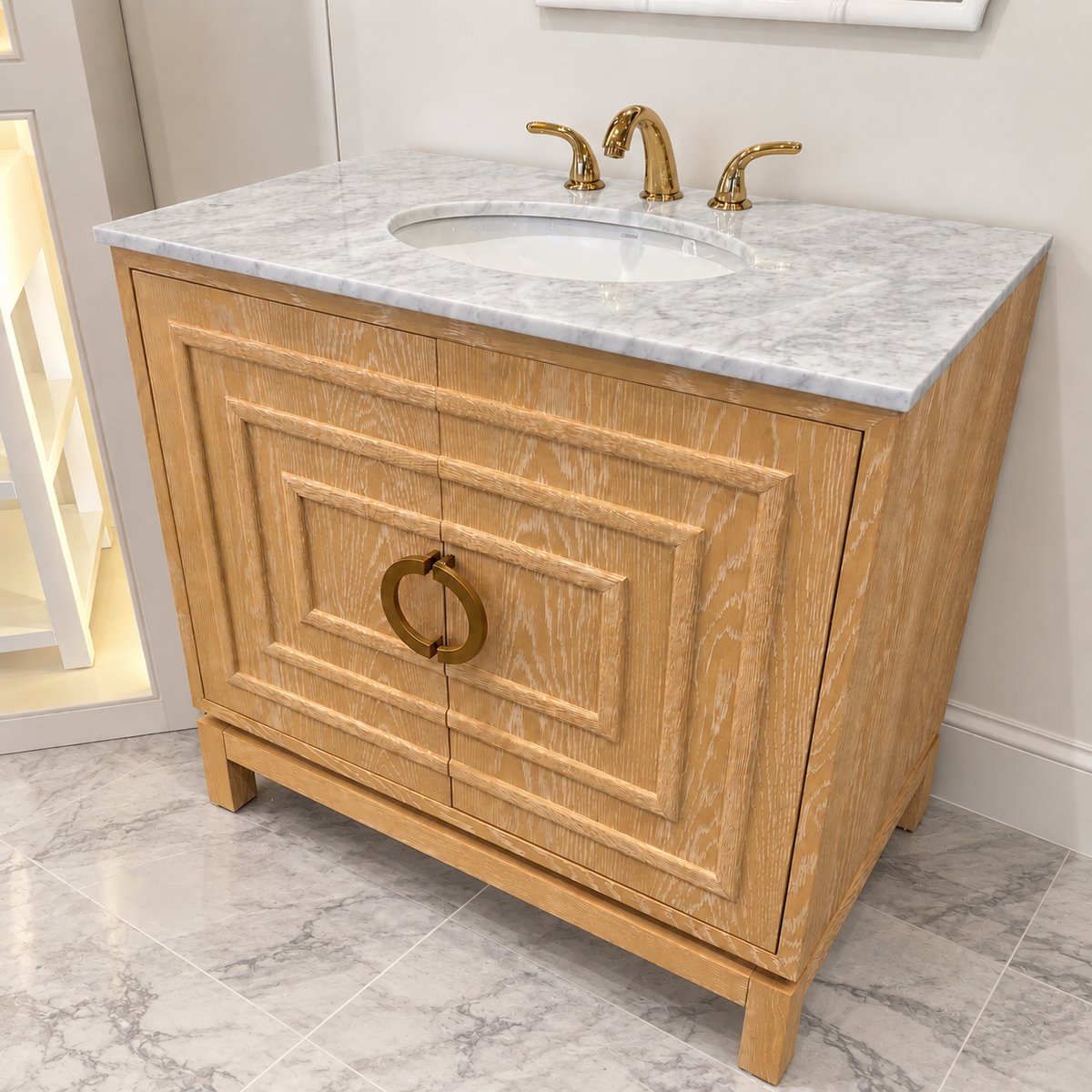 Cerused oak bathroom vanity with geometric panel detailing, brass ring hardware, white marble countertop, and gold faucet on cool marble tile flooring.