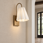 A modern wall sconce light fixture with a warm brass-finished metal frame and a pleated white glass shade, mounted on a textured beige wall in an interior hallway.