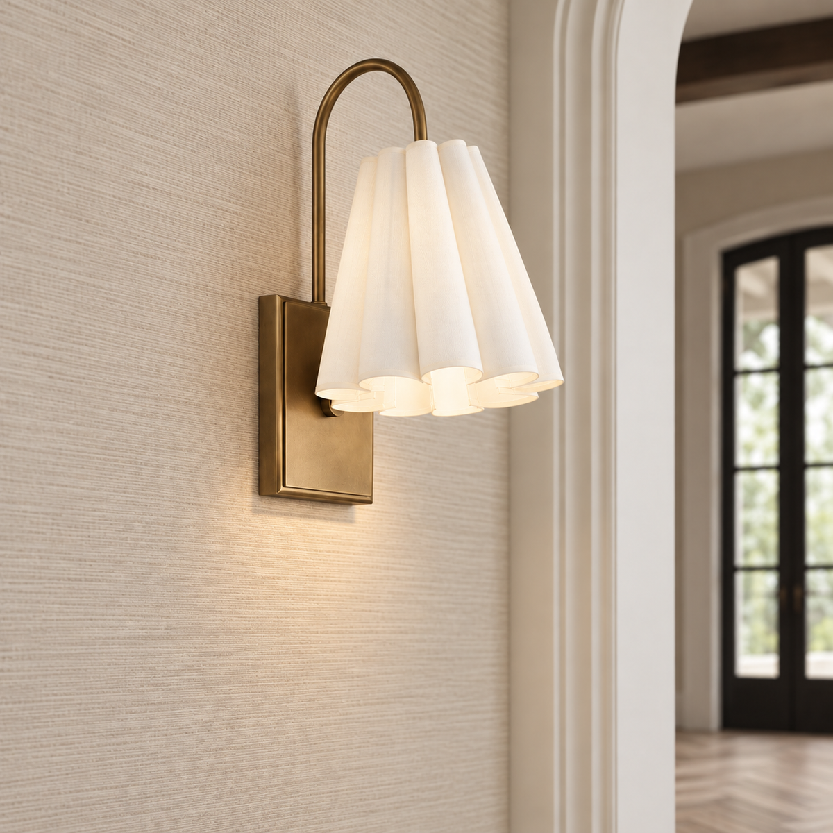 A modern wall sconce light fixture with a warm brass-finished metal frame and a pleated white glass shade, mounted on a textured beige wall in an interior hallway.