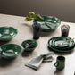 Green ceramic tableware set on a beige surface with a plant and glasses.