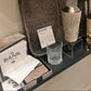 A hand is shown placing a large gray felt tray onto a shelf in a showroom. The tray is displayed with various bar accessories, including a textured cocktail shaker, a small textured glass, a gold rabbit figurine, and other decorative items.