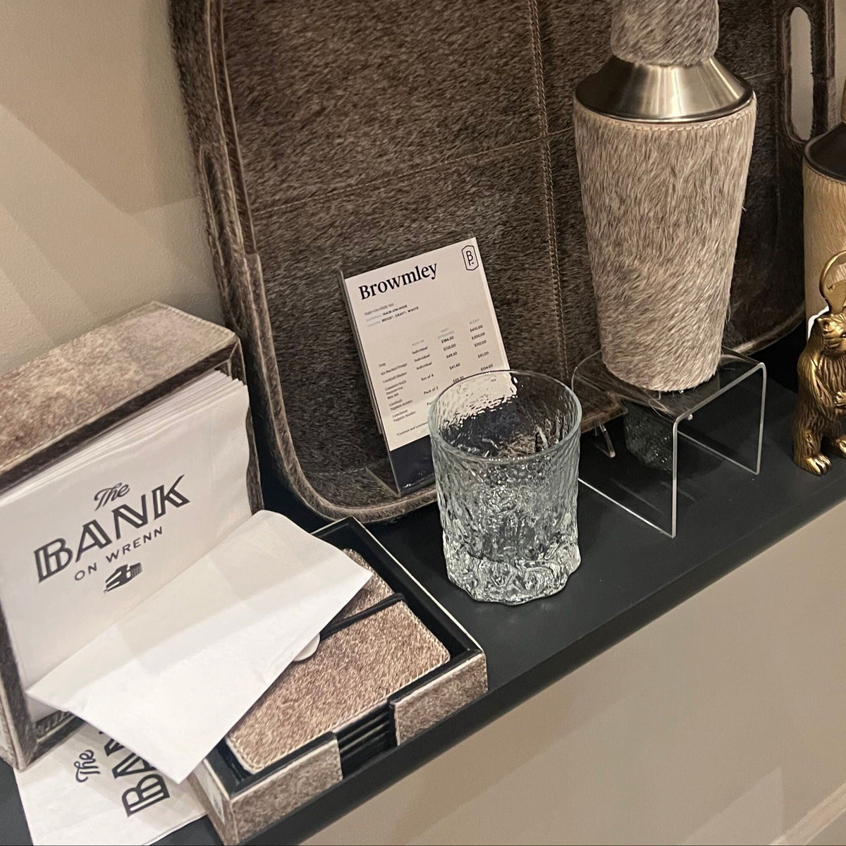 A hand is shown placing a large gray felt tray onto a shelf in a showroom. The tray is displayed with various bar accessories, including a textured cocktail shaker, a small textured glass, a gold rabbit figurine, and other decorative items.