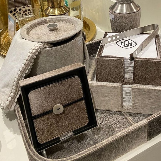 A styled home decor display featuring a large rectangular tray with a textured taupe and gray border, holding a small black-and-beige textured clutch, a folded napkin holder with a silver metal logo, and a patterned tissue box. The tray is surrounded by other decorative items on a white retail display table.