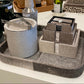 A styled home decor and bar accessory set displayed on a white table, featuring a gray and brown textured tray with a cylindrical bucket container, a cocktail shaker, and a tiered jewelry organizer.