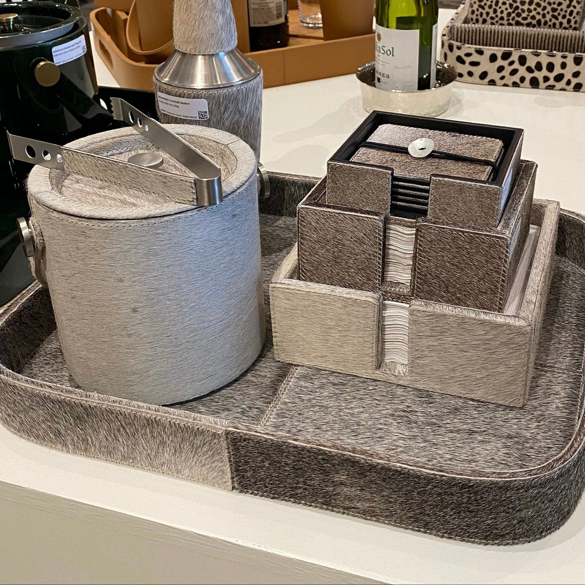 A styled home decor and bar accessory set displayed on a white table, featuring a gray and brown textured tray with a cylindrical bucket container, a cocktail shaker, and a tiered jewelry organizer.