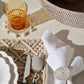 A styled tabletop setting with a white cloth napkin secured by a round woven ring, a silver spoon and knife on a textured beige placemat, a white plate edge, an amber glass tumbler on a pom-pom coaster, and part of a woven basket in the background.