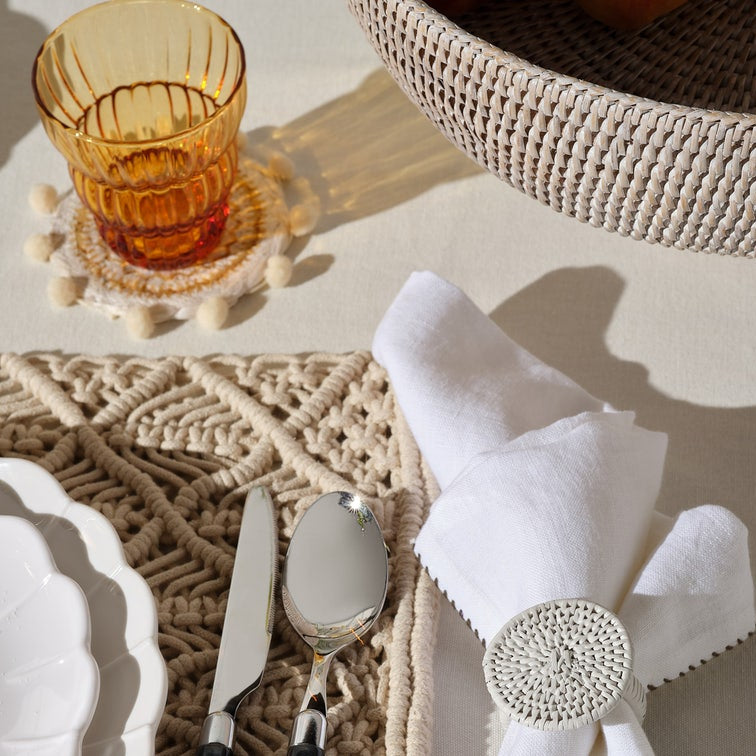 A styled tabletop setting with a white cloth napkin secured by a round woven ring, a silver spoon and knife on a textured beige placemat, a white plate edge, an amber glass tumbler on a pom-pom coaster, and part of a woven basket in the background.