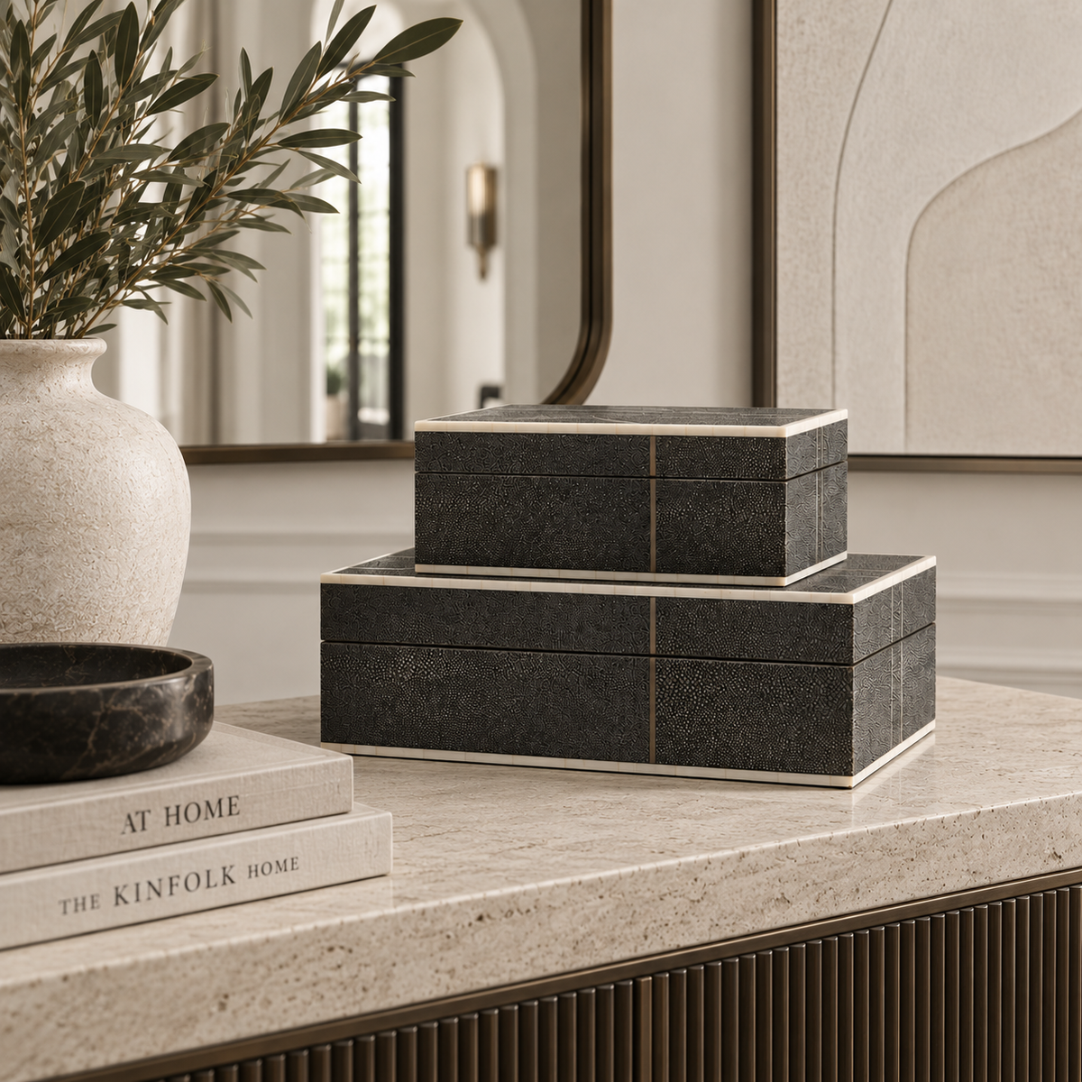 Two stacked rectangular black textured boxes with cream-colored edges sit on a light stone console table in a styled interior setting.