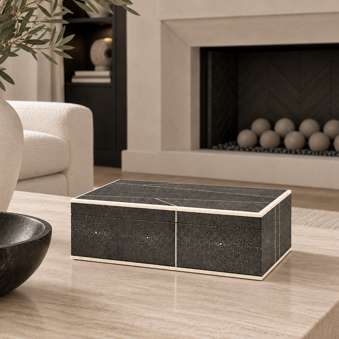 A rectangular black textured storage box with cream-colored trim sits on a light beige stone table in a modern living room setting with a fireplace in the background.