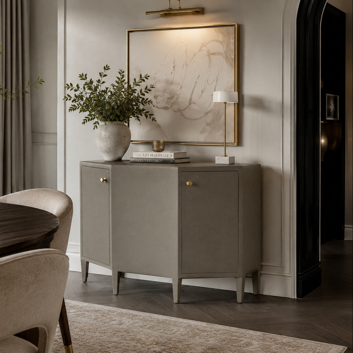A modern gray sideboard cabinet in a styled dining room setting, decorated with a white ceramic vase of greenery, books, and a small table lamp, beneath a gold-framed abstract art print.