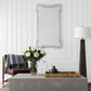 A rectangular wall mirror with a silver-toned geometric frame hangs on a white paneled wall above a gray console table. The scene includes a brown leather chair with a blue throw, a vase of white flowers, a bamboo tray, and a tall black floor lamp.