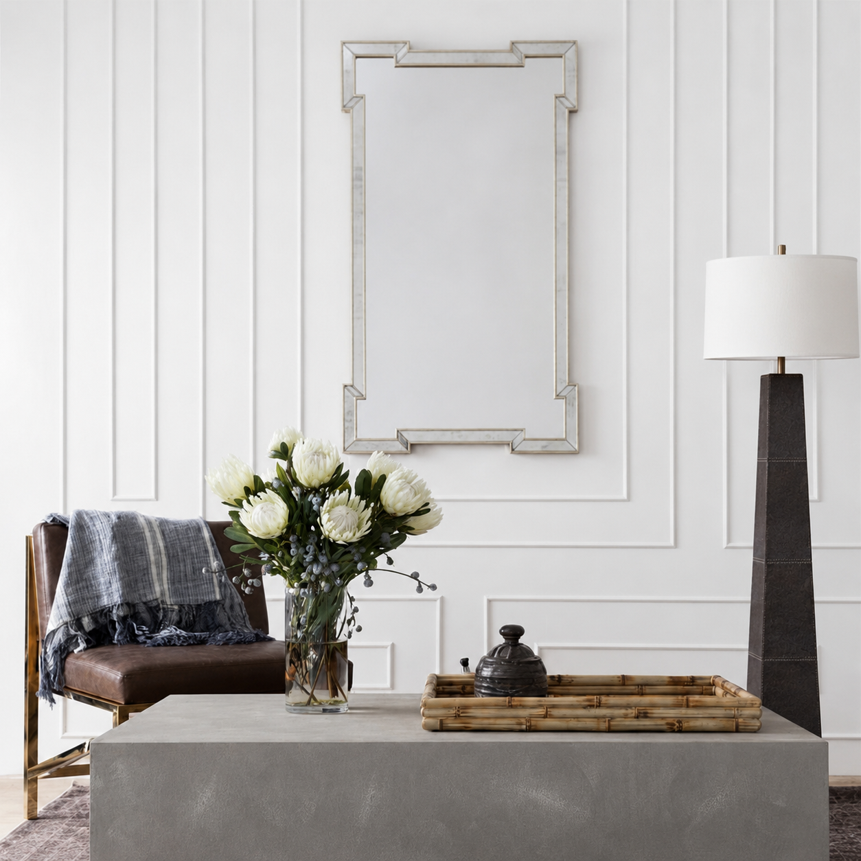 A rectangular wall mirror with a silver-toned geometric frame hangs on a white paneled wall above a gray console table. The scene includes a brown leather chair with a blue throw, a vase of white flowers, a bamboo tray, and a tall black floor lamp.