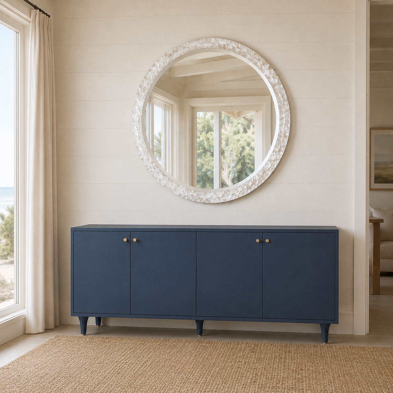 A navy blue wooden credenza with small brass knobs, positioned below a large round white mirror on a shiplap wall in a bright, coastal-style room.