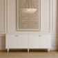 A modern pendant light with a frosted white bowl shade and bronze chain hardware hangs in front of a large framed textured artwork above a white sideboard cabinet in a classic cream paneled room.