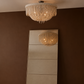 A large rectangular floor mirror with a thin metallic frame leans against a dark brown wall on a light wood floor. The mirror reflects a hanging circular chandelier with beige fringe and warm glowing bulbs.