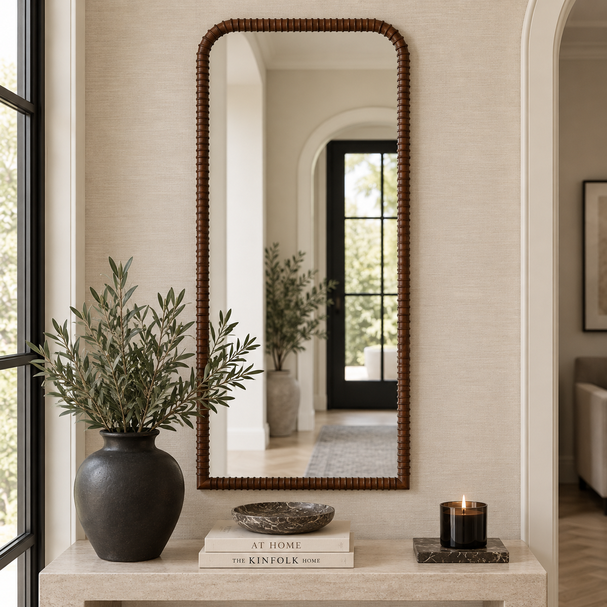 A tall rectangular mirror with a dark brown beaded frame hangs on a light beige textured wall above a console table styled with a dark vase of greenery, stacked books, and a small candle.