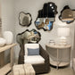 A styled interior scene featuring a dark woven rattan armchair with white cushions, a matching ottoman, a curved beige side table with a white table lamp, and several organic-shaped dark-framed mirrors on a light gray wall.