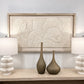 A large rectangular framed wall art piece with a textured, embossed floral or sunburst design in warm off-white tones, displayed above a console table with two white ribbed table lamps and two brown vases.